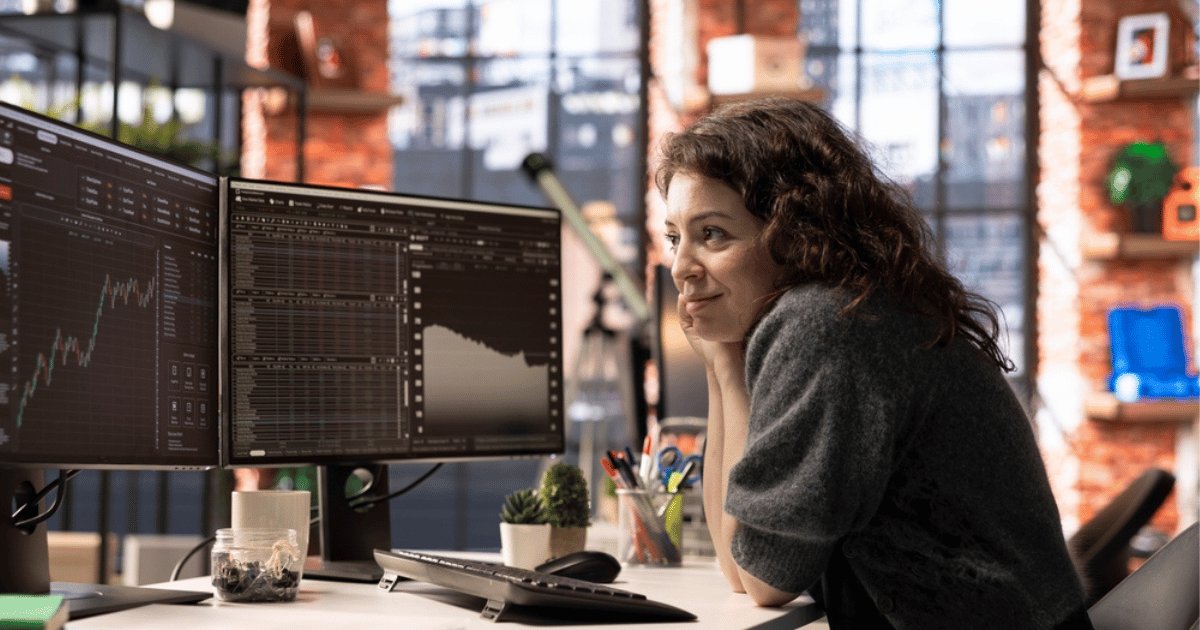Woman at Desk Contemplating Two Screens With Charts